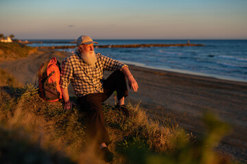Elderly Man with Long White Beard Sitting on a Coastal Cliff at Sunset, Reflecting on the Ocean View