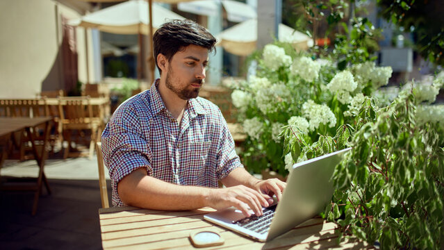 Business man working on laptop sitting at cafe table outdoors. Concept of remote work in public space, freelance productivity, digital entrepreneurship, modern corporate lifestyle