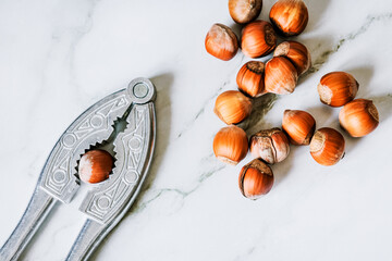 Close-up flat-lay: vintage metal nutcracker gripping a hazelnut, pile of filberts on white marble,...