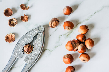 Close-up flat-lay: vintage metal nutcracker gripping a hazelnut, pile of filberts on white marble,...
