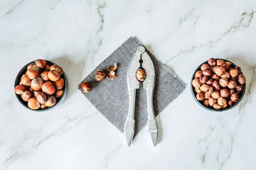 Symmetrical flat-lay: twin bowls of whole and shelled hazelnuts flank ornate metal nutcracker on...