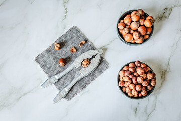 Overhead layout of hazelnuts in two ceramic bowls beside vintage nutcracker on grey woven cloth...