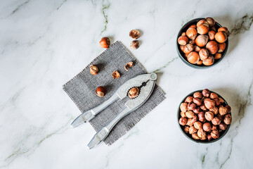 Overhead layout of hazelnuts in two ceramic bowls beside vintage nutcracker on grey woven cloth...