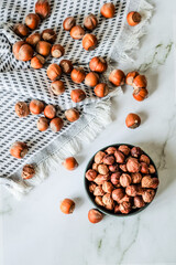 Top-view of scattered hazelnuts on textured cotton kitchen towel with bowl of shelled nuts on white...