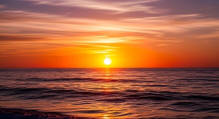 The sun sets over the ocean, casting a brilliant orange and red glow across the water and sky, creating a beautiful and peaceful natural scene.

