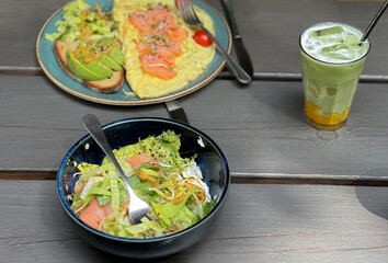 Two plates of avocado and red fish dishes served on a wooden table with a glass of matcha mango served with a metal straw. Healthy breakfast al fresco in natural light.


