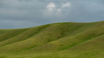 Rolling green hills under a cloudy sky create a serene landscape, with gentle slopes and a solitary figure in the distance, embodying tranquility and nature's beauty