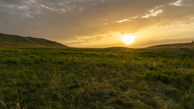 Expansive green landscape under a dramatic sky, with rolling hills illuminated by a warm sunset, creating a serene and tranquil atmosphere in nature's beauty