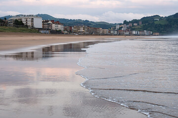 Waves gently lap the shore in Zarautz, capturing reflections of coastal buildings at dawn