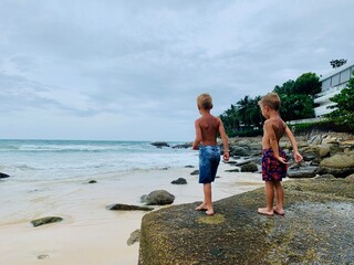 Two preschool boys enjoy a sunny day on a tropical beach. Sandy shore, clear water, and large rocks...