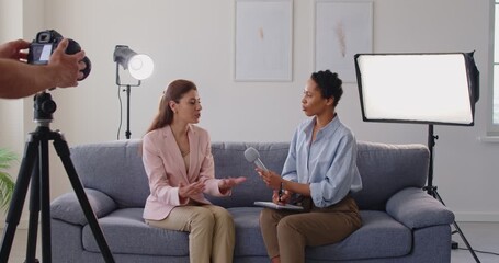 Media interview, TV talk show recording, studio dialogue of business lady and journalist with microphone sitting on sofa. Guest talking about story with gestures, reporter listening, cameraman filming