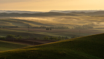 Fototapeta premium Long mountains. Serene landscape at dawn, showcasing rolling hills covered in lush greenery, with soft mist rising above fields, creating a tranquil and picturesque rural scene