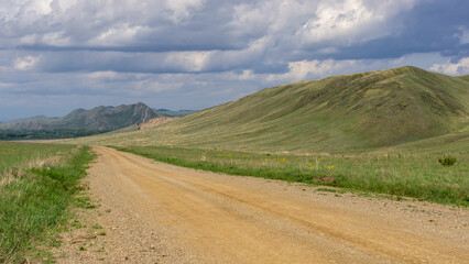 Long mountains. Scenic dirt road winding through lush green hills under a dramatic sky filled with clouds, inviting exploration and adventure in a tranquil natural landscape