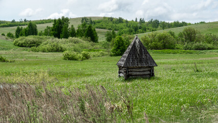 Obraz premium Rustic wooden structure with a triangular roof stands alone in a lush green meadow, surrounded by rolling hills and trees under a bright cloudy sky, evoking tranquility and solitude