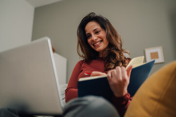 Smiling woman taking notes while working from home using laptop and agenda