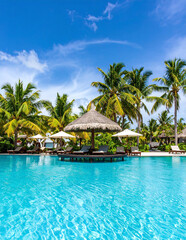 Beautiful tropical resort swimming pool with crystal-clear turquoise water sparkling under bright midday sun surrounded by palm trees and beach umbrellas