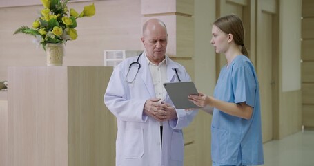 senior caucasian male doctor discussing diagnosis with young caucasian female nurse while reviewing patient data on digital tablet near nurse counter inside hospital showing clinical teamwork - Powered by Adobe