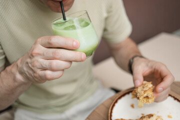 Close-up of a mid adult man sitting outdoors in summer drinking an iced matcha latte and eating a freshly baked croissant.