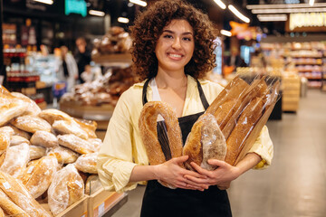 Standing in the supermarket and smiling. Female worker in apron is with bakery in hands