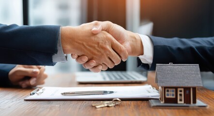 A real estate agent and customer shaking hands over the table with a house model and keys on the desk