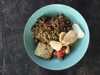 Top view of Indonesian nasi goreng served with tempe goreng, krupuk crackers, and spicy sambal on a blue plate. Rustic wooden table background. Authentic Asian street food concept.