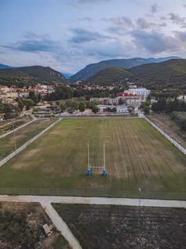 Aerial view of a vibrant green sports field with blue goalposts nestled between terracotta rooftops and verdant hills under a pastel sky, Prades, Occitanie, France.