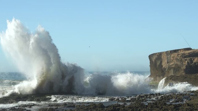 SLOW MOTION: Massive wave crashes with great force against a rocky cliffside and the surrounding shoreline rocks. White spray of the wave contrasts sharply with the blue sky. Raw power of the ocean.