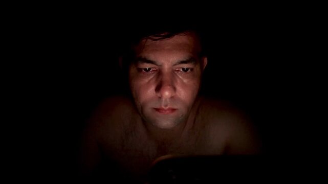 Close up, a tired man in bed scrolls endlessly on his mobile phone, struggling with insomnia, anxiety and overthinking as he watches social media late into the night