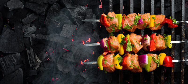 Raw Chicken Skewers with Vegetables on a grill grate, over hot charcoal. Top view. On a black textured background. Free space for text.