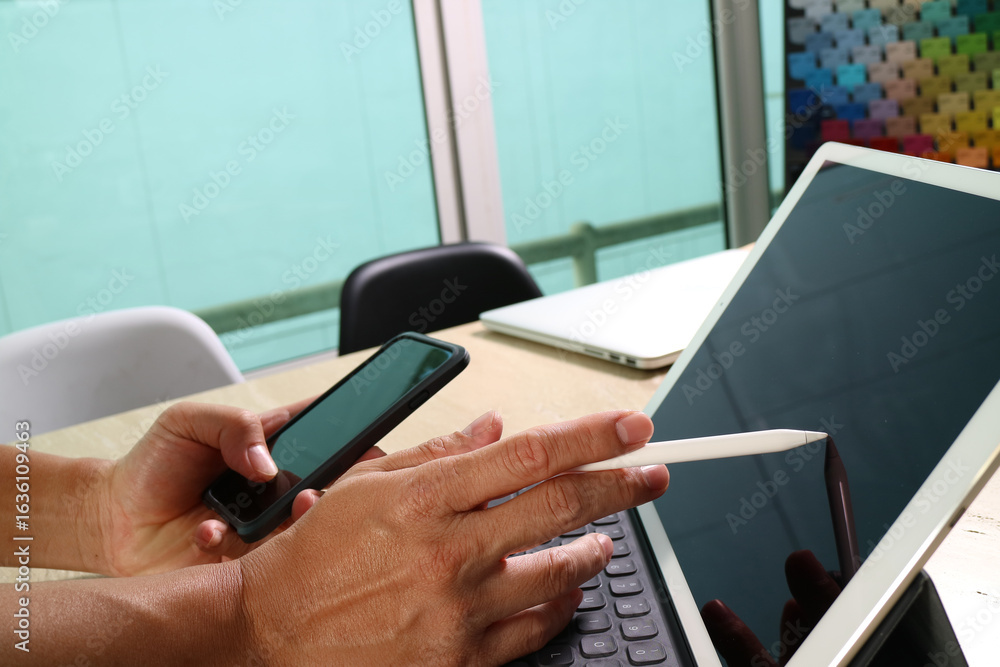 Poster creative designer using stylus on tablet while holding smartphone at workspace