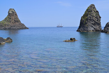Faraglioni of Acitrezza with tall ship sailing in Ionian sea. Volcanic sea stacks and clear water with a historic tall ship sailing the horizon.