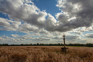Solitary stone cross in golden wheat field beneath dramatic sky with heavy clouds. Peaceful and...