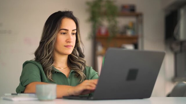 Frustrated Latin businesswoman in green shirt gesturing with hand while looking at laptop with concerned expression in home office setting during challenging virtual meeting or technical problem