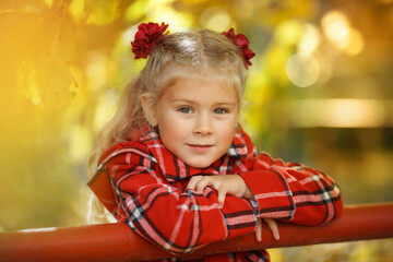 Portrait of a blonde girl in a red plaid coat leaning on a red metal railing with her elbows,...