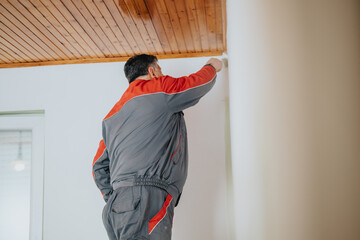 A professional worker wearing a gray and red uniform carefully painting a white interior wall,...