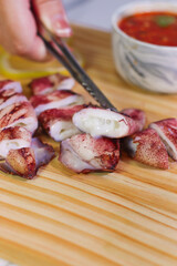 Hand Holding Metal Tongs Picking Up Grilled Squid Piece on Wooden Board with Dipping Sauce and Lime