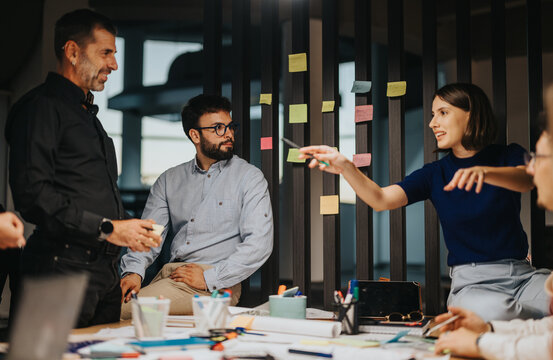 Business team discussing ideas in a modern workspace, engaging in creative collaboration. Notes are pinned to a board, and documents spread across the table highlight their focus and teamwork.