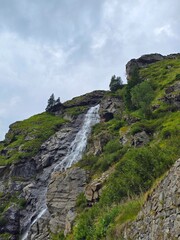 View of the Capra Waterfall from the Transfagarasan Road on a summer day, with green grass, rocks and a cloudy sky. Arges, Romania