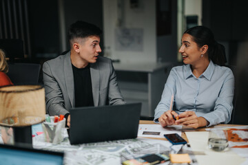 Two colleagues engage in a late-night discussion at the office, collaborating on business strategies. Papers and laptops are spread across the table, showcasing teamwork and dedication.