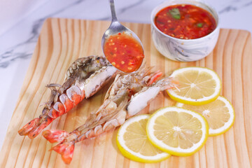 Person Pouring Seafood Dipping Sauce on Grilled River Prawn Head on Pine Cutting Board with Lemon Slices