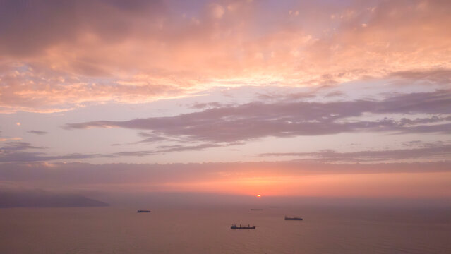 Tranquil sunset over the Mediterranean Sea with pastel clouds in orange, pink, and lavender hues, and cargo ships floating calmly on the water under a glowing horizon.