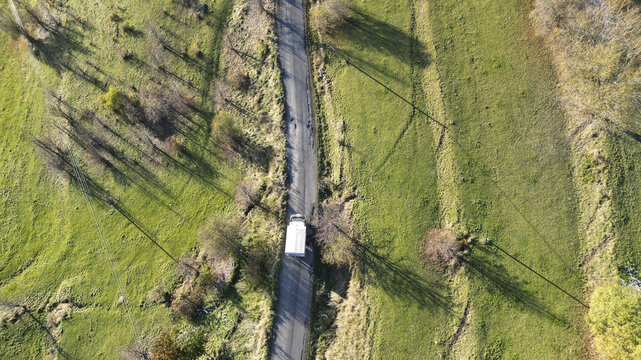Aerial view of a lone white van navigates a narrow asphalt road cutting through vibrant green fields casting long shadows, Posof, Ardahan, T&Atilde;&frac14;rkiye.