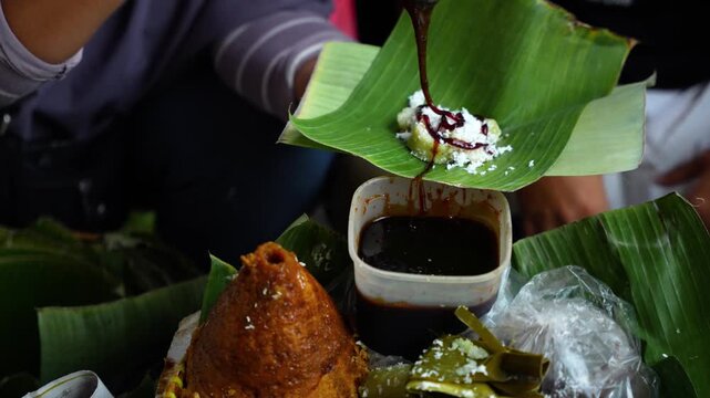 Handheld close-up of a street vendor finishing jajan pasar: palm sugar syrup poured over a banana leaf parcel topped with grated coconut, completing a classic Indonesian market dessert.