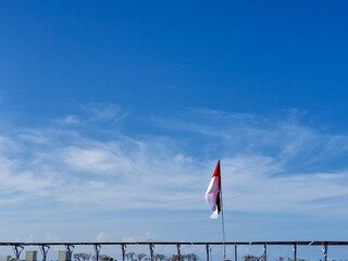 A lone red and white flag stands proudly against a vast, clear blue sky with soft, wispy clouds....