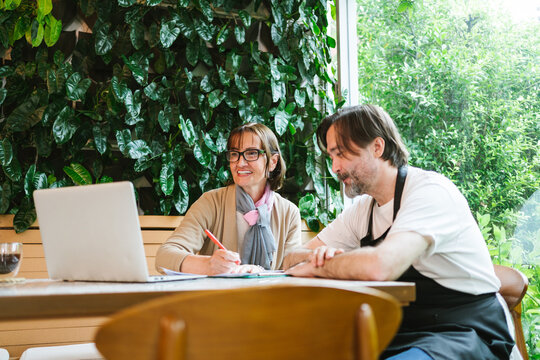 Small business owner and financial advisor smiling while reviewing documents on a laptop in a cozy cafe, highlighting diverse collaboration, business growth, and planning in a relaxed setting.