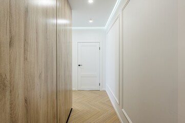 Bright hallway with light wood flooring, a large wood cabinet, and white walls with decorative molding. A white door is centered at the end of the hall