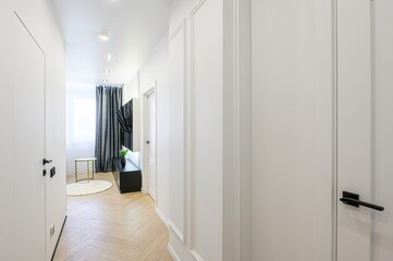 Bright hallway with white walls, doors, and herringbone wood floors. A glimpse of a cozy room with...