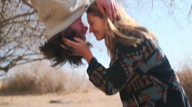 A cheerful young man hangs upside down from a tree while a woman with long blonde hair in a patterned jacket cups his head. They smile, nuzzling noses against bare trees and bright spring sunshine.