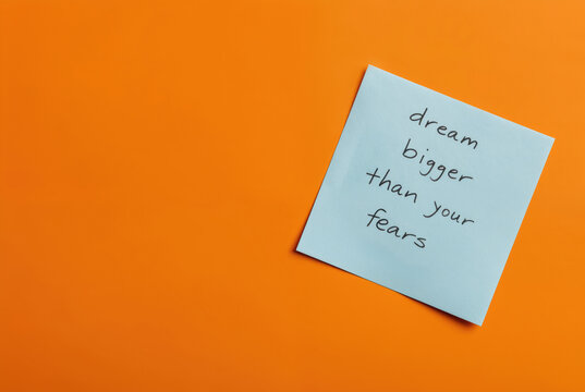 Note reminding to dream bigger than fears, handwritten on blue paper over vibrant orange background. Motivational affirmation, self-improvement message for overcoming obstacles. Inspirational quote