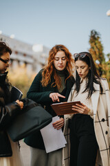 Three young women collaborate and review tasks on a tablet, discussing work outside under a clear sky. Their expressions emphasize communication and planning in a casual yet professional environment. © qunica.com